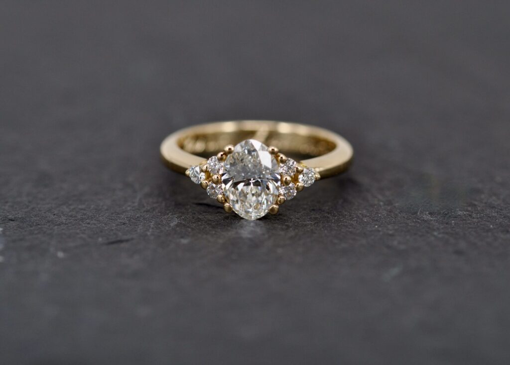 Handgefertigter Verlobungsring aus 750 Gelbgold von Goldschmiede Stephanie Berger mit ovalem Hauptdiamanten und mehreren funkelnden Brillanten.